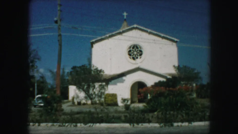 1959 Kansas City 8mm view of a white church front with a cross, round window, arched entrance, roadside greenery, and a car partly visible at left.