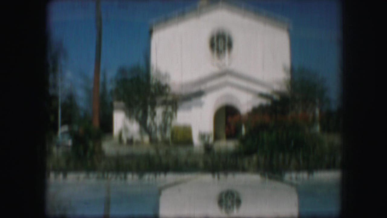 White church exterior with rose window, arched entry, shrubs, and blue sky.