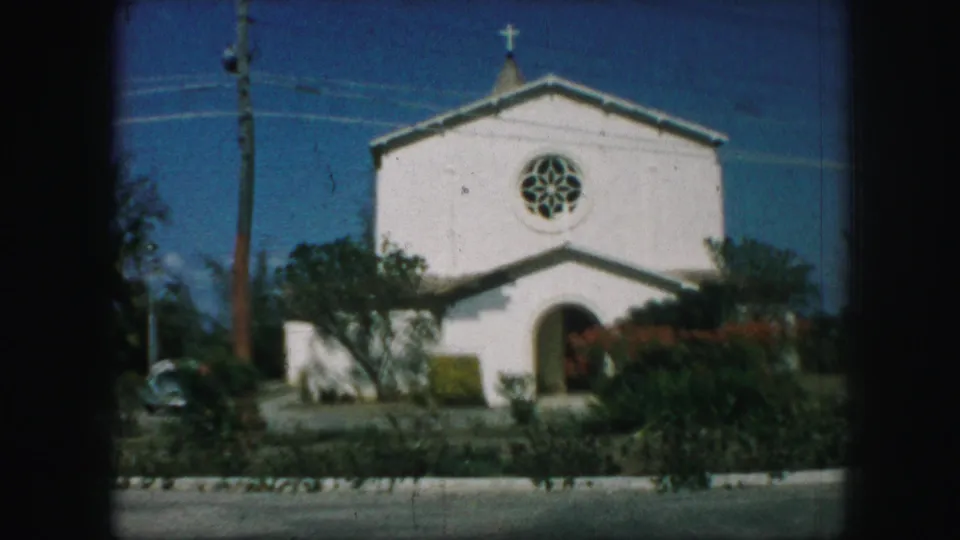 1959 Kansas City 8mm frame centered on a white church with rose window, cross, arched entry, roadside foreground, and shrubs.