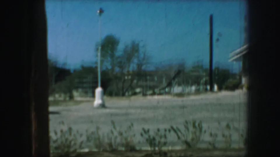 1959 Kansas City home-movie frame showing surrounding land near the church, with a pole, sparse trees, low structures, and film border.
