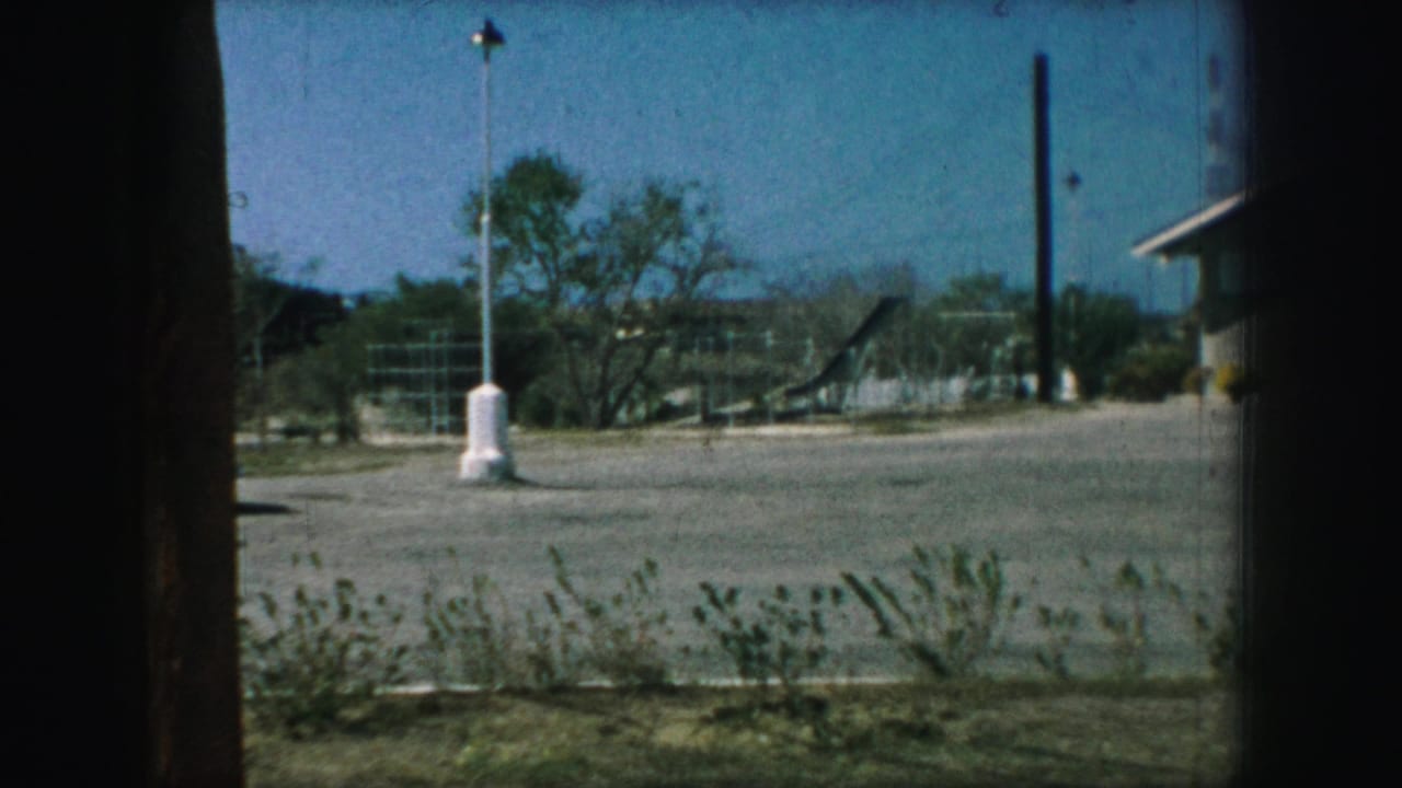 View through a dark window frame toward a gravel lot, lamppost, trees, and part of a low building.