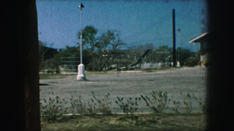 1959 Kansas City 8mm frame looking through a window or doorway toward a gravel lot, lamppost, trees, and a low modern building edge.