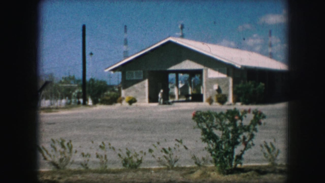 1959 Kansas City exterior view of a low sloped-roof building with a glass entrance, gravel foreground, bushes, and utility poles.