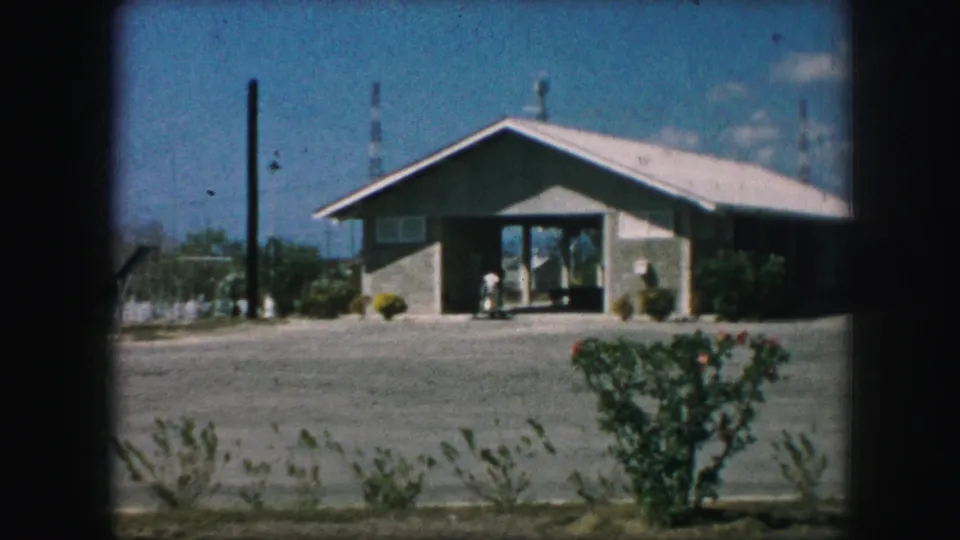1959 Kansas City 8mm frame of the low building and entrance seen through dark window edges, with gravel and bushes in front.