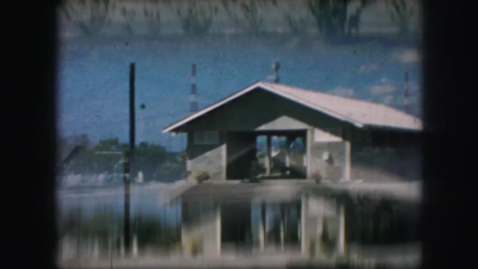 End frame from 1959 Kansas City footage showing a blurred handheld view of the building entrance through dark framing, with gravel and bushes.