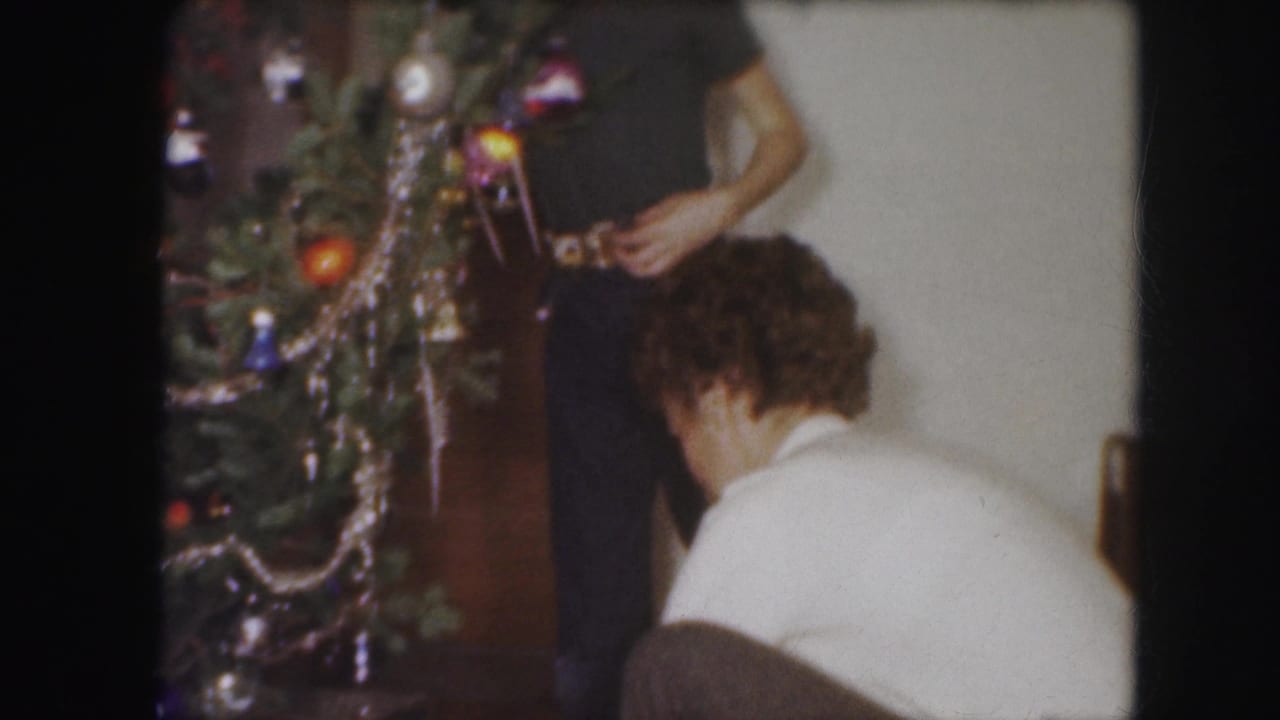 1959 Kansas City home-movie frame showing adults near a Christmas tree with ornaments and tinsel in a private interior.