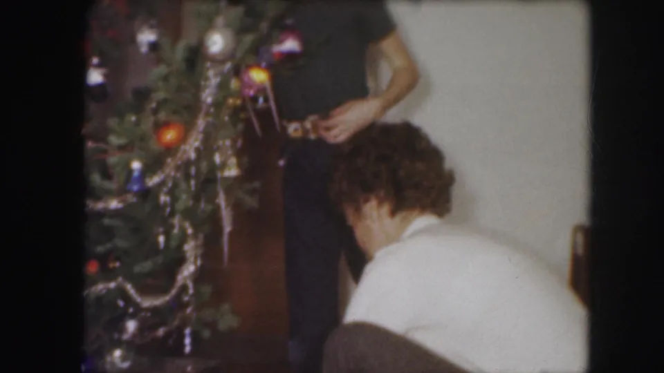 1959 Kansas City home-movie frame showing adults near a Christmas tree with ornaments and tinsel in a private interior.
