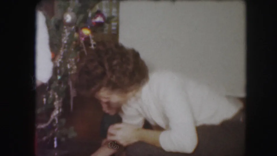 1959 Kansas City home-movie frame showing a woman leaning near a decorated Christmas tree inside a private room.