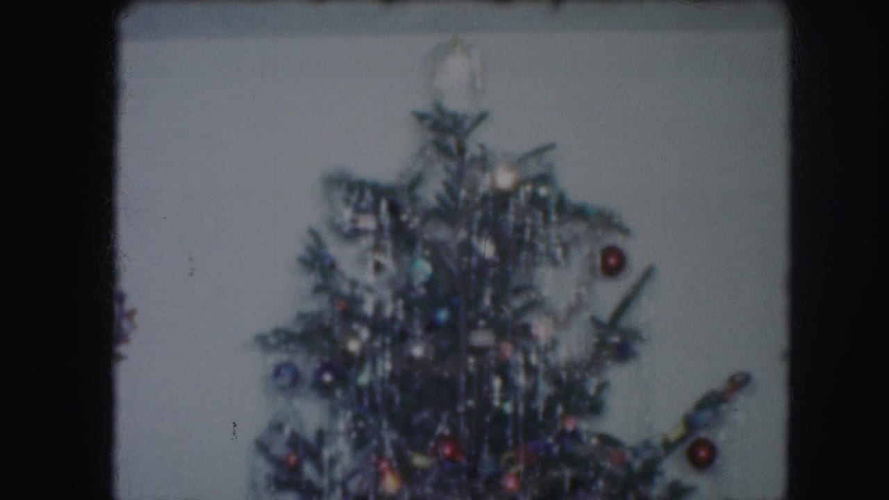 Top portion of a tinsel-covered Christmas tree with ornaments against a pale wall.