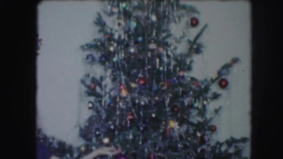 Mid-pan view of the Christmas tree with a partial person or arm entering near the lower left in archival 8mm footage.