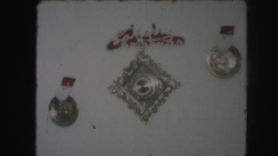 A Kansas City 1959 home-movie frame showing silver medals and a decorative diamond-shaped emblem beneath red Arabic script.