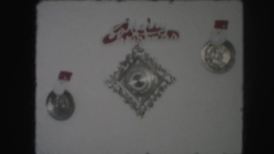 1959 8mm frame showing a home wall display of silver medals, red Arabic script, and a diamond-shaped decorative emblem.