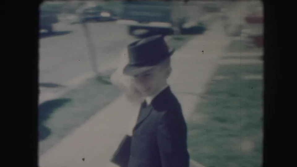 A closer 8mm frame shows the boy's dark suit and hat as he walks beside the girl in white, with yard and cars behind them.