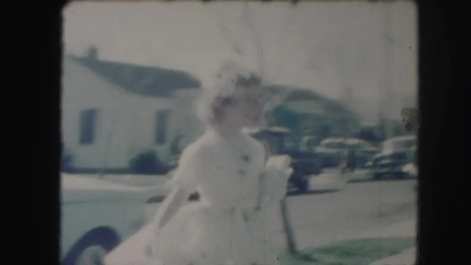 The girl in a white dress is seen near the end of the clip, with a suburban house and mid-century cars in the background.