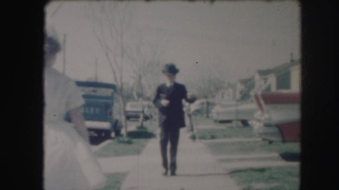 A man in a dark suit and hat walks along a residential sidewalk with parked cars and modest houses in the background.