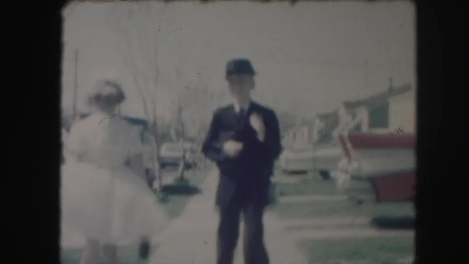 A woman in a light dress passes near a man in a dark suit on a suburban sidewalk lined with cars and houses.