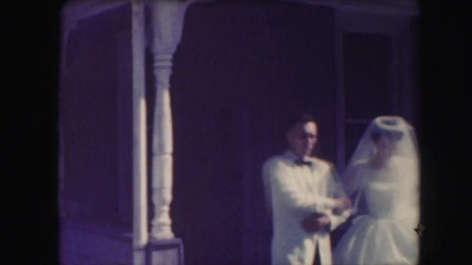 1959 Kansas City home-movie still of the groom in a light jacket and bride in veil on a shaded porch.