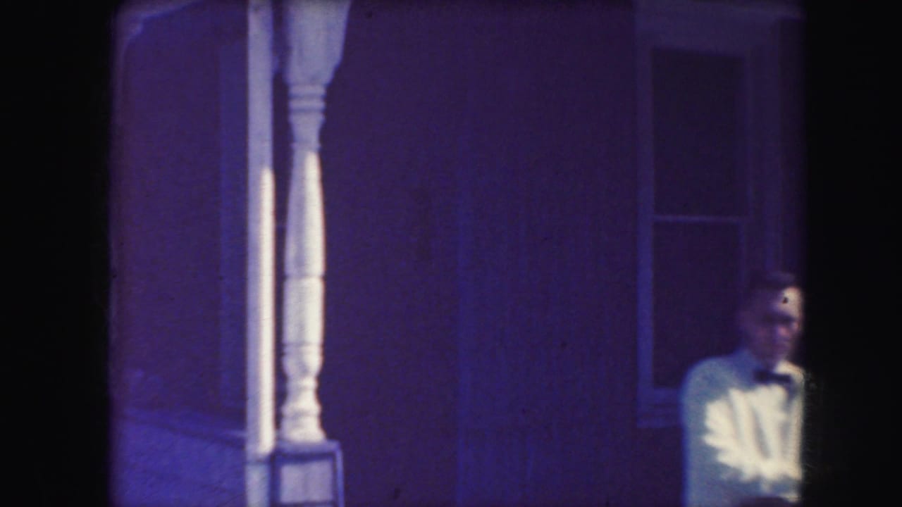 Man in formal clothing stands near a house porch.