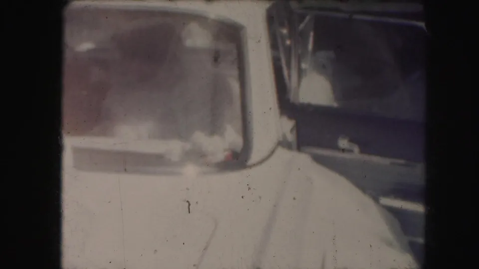 The bride's white dress and veil fill the foreground as she moves near the open rear door of the midcentury car.