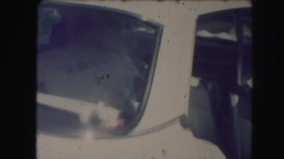 The bride appears partly inside or beside the open rear car door, with the car window and door frame prominent in the home-movie frame.