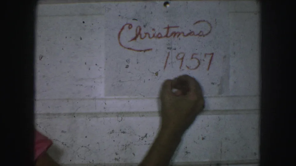 Christmas 1957 is visible in red marker on a white paneled wall as the hand finishes the written date cue.
