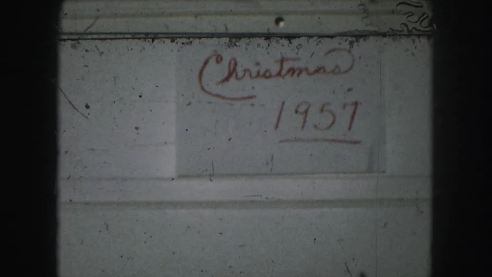 A late-1950s 8mm frame shows the red handwritten title “Christmas 1957” on a pale wall or board, with grain and edge shadows.