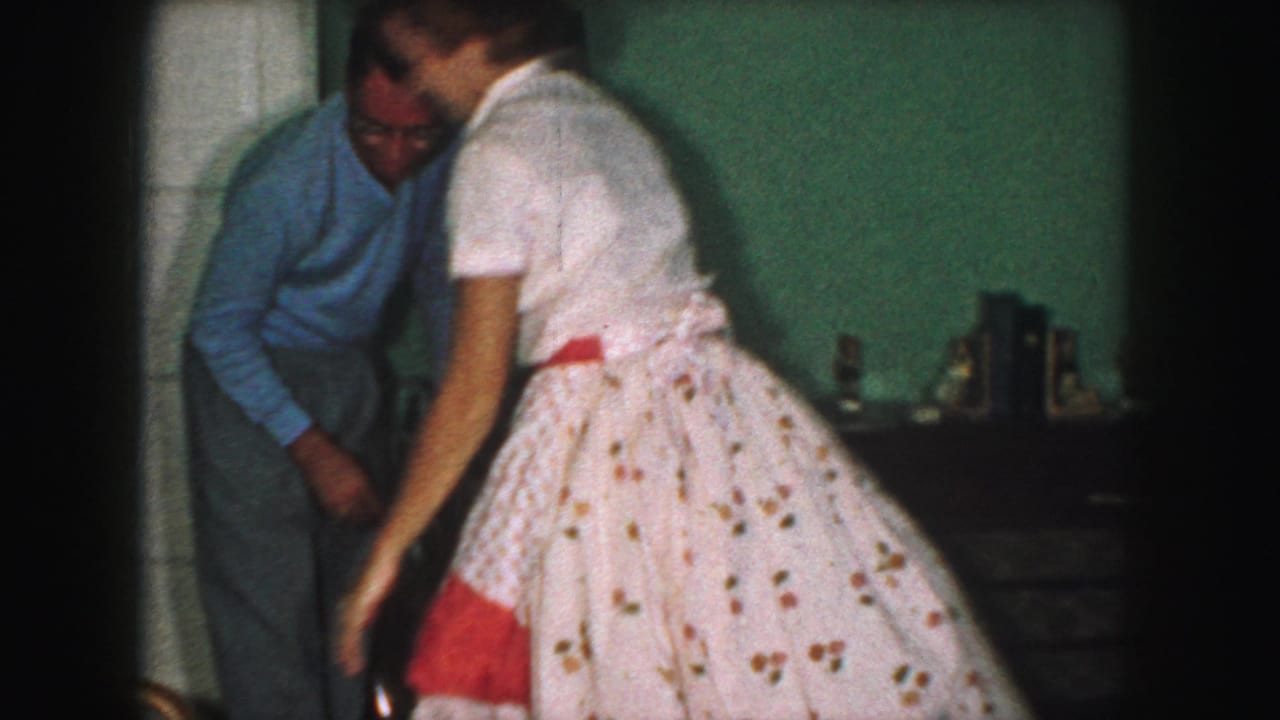 In a 1959 Kansas City home-movie frame, a woman in a white dress with red sash stands near a man in a blue shirt inside a private room.