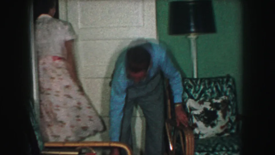 The woman in a white floral dress moves toward a door as the man in a blue shirt bends beside a patterned chair in the Kansas City home interior.