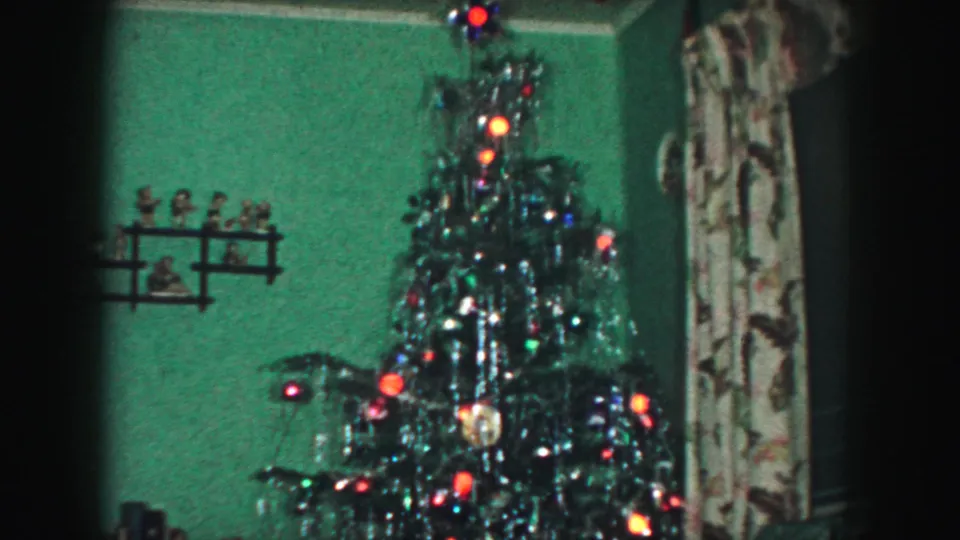 A decorated Christmas tree with tinsel, ornaments, and colored lights stands against a green wall in the 1959 home-movie interior.
