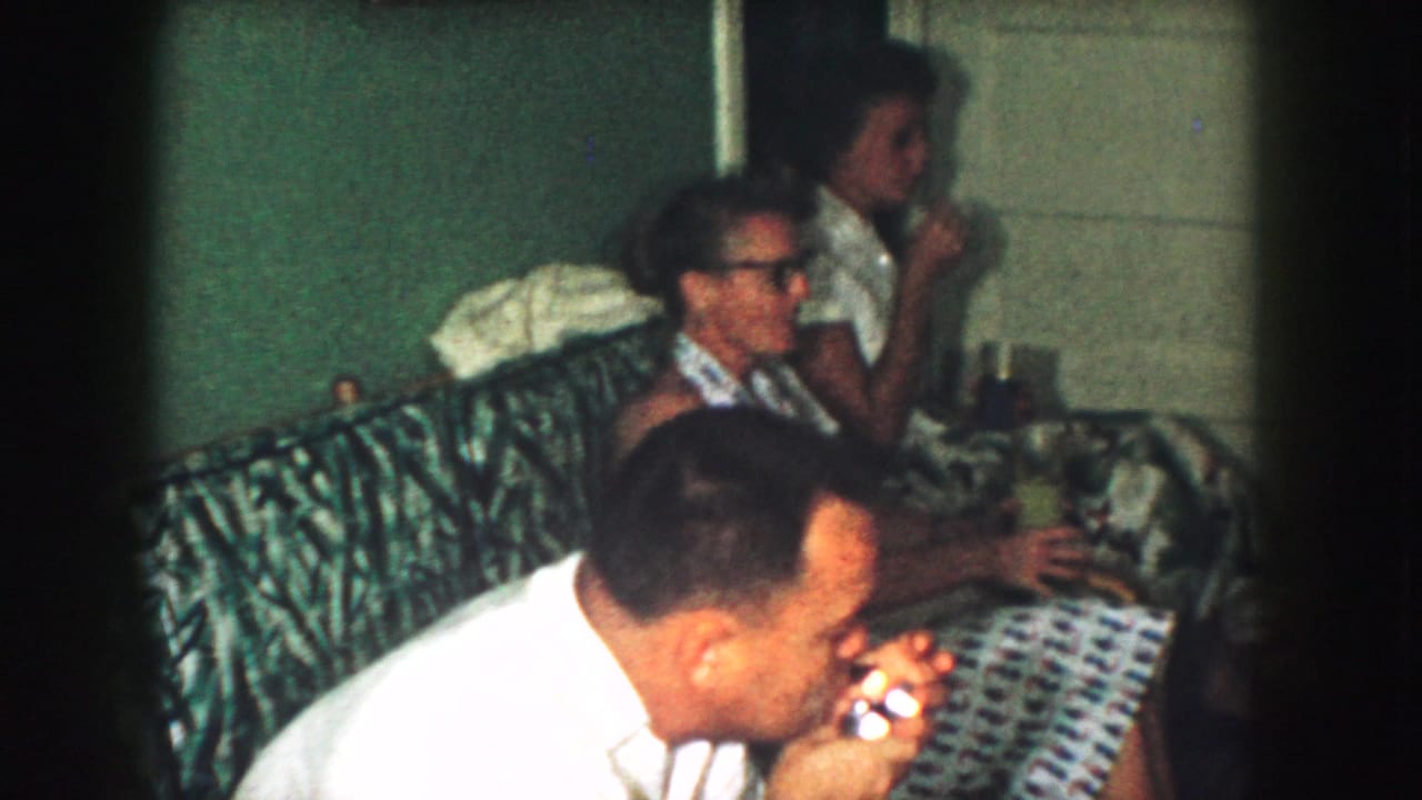 1959 Kansas City home-movie frame with adults gathered on a patterned couch, one man leaning forward and others holding drinks.