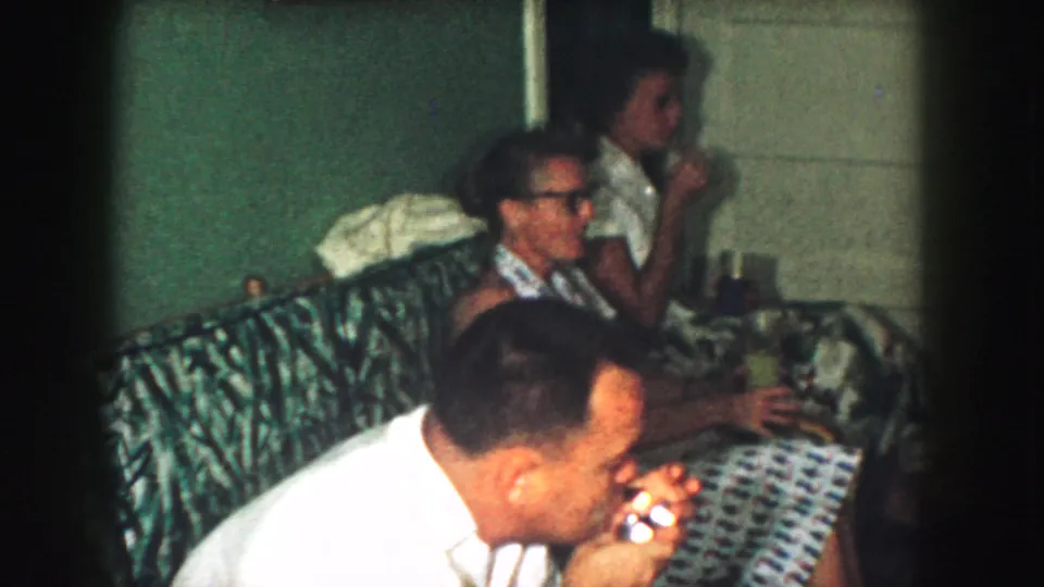 1959 Kansas City home-movie frame with adults gathered on a patterned couch, one man leaning forward and others holding drinks.