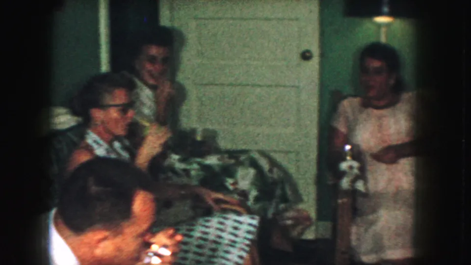 1959 Kansas City home-movie frame showing adults talking in a living room with bottles, a patterned couch, and a black lampshade nearby.