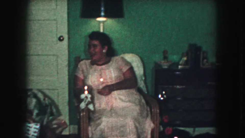 1959 Kansas City home-movie frame of a woman seated in a chair beside a small dresser, bottles, and a black floor lamp.