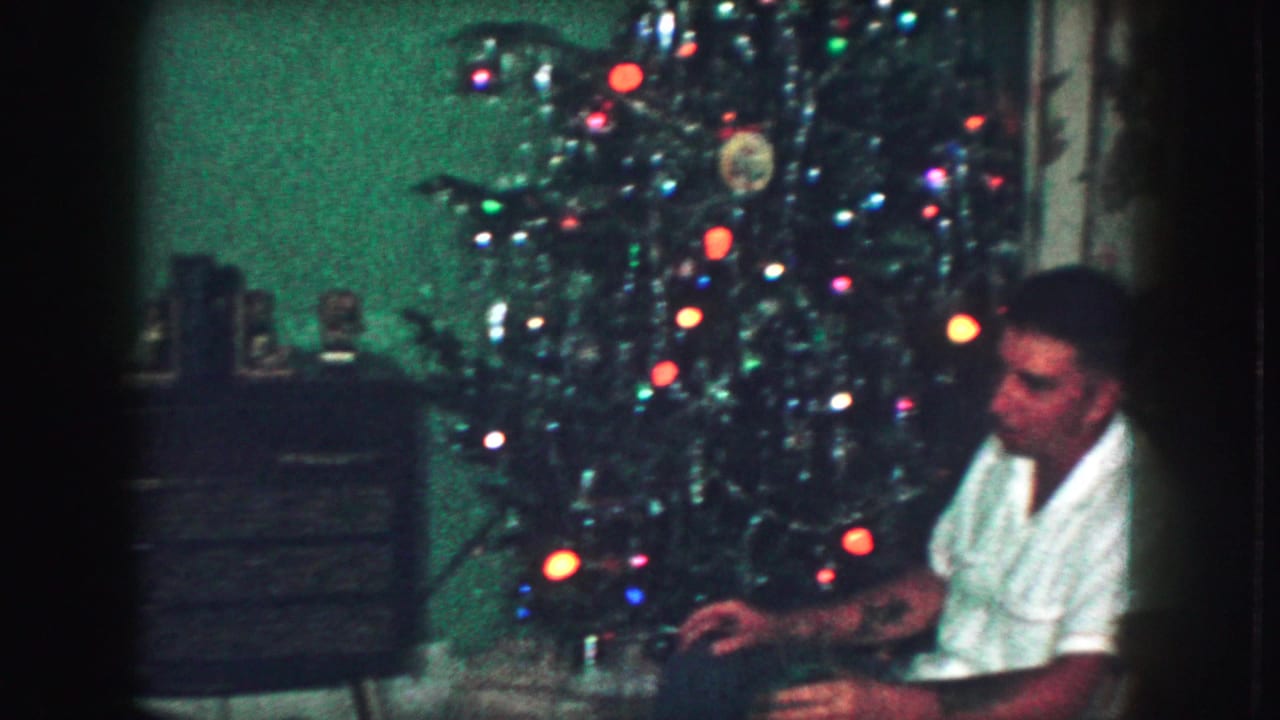 In a 1959 Kansas City home-movie frame, a man in a white shirt sits beside a lit Christmas tree in a private living room.