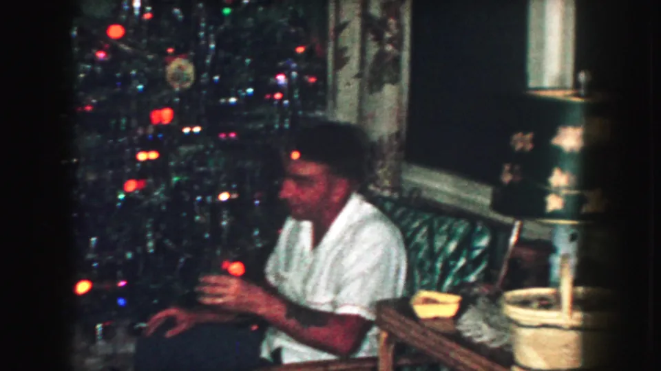 A man sits with a drink near the Christmas tree, with a lamp, side table, ashtray, and curtains visible in the living room.