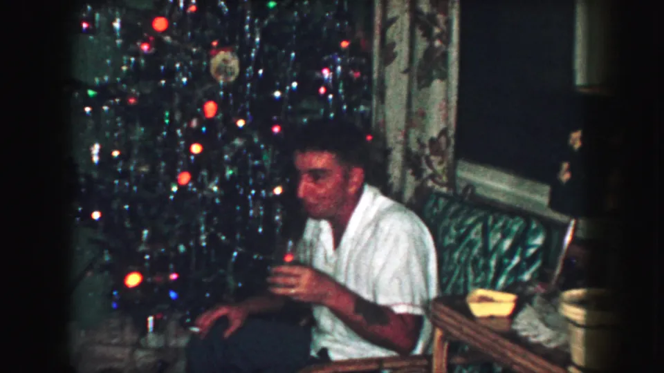 In a 1959 Kansas City living room, a man in a white shirt holds a drink beside a decorated Christmas tree and tabletop lamp.