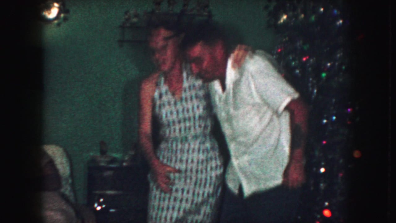 Adult couple dancing near a lit Christmas tree in a dim living room.