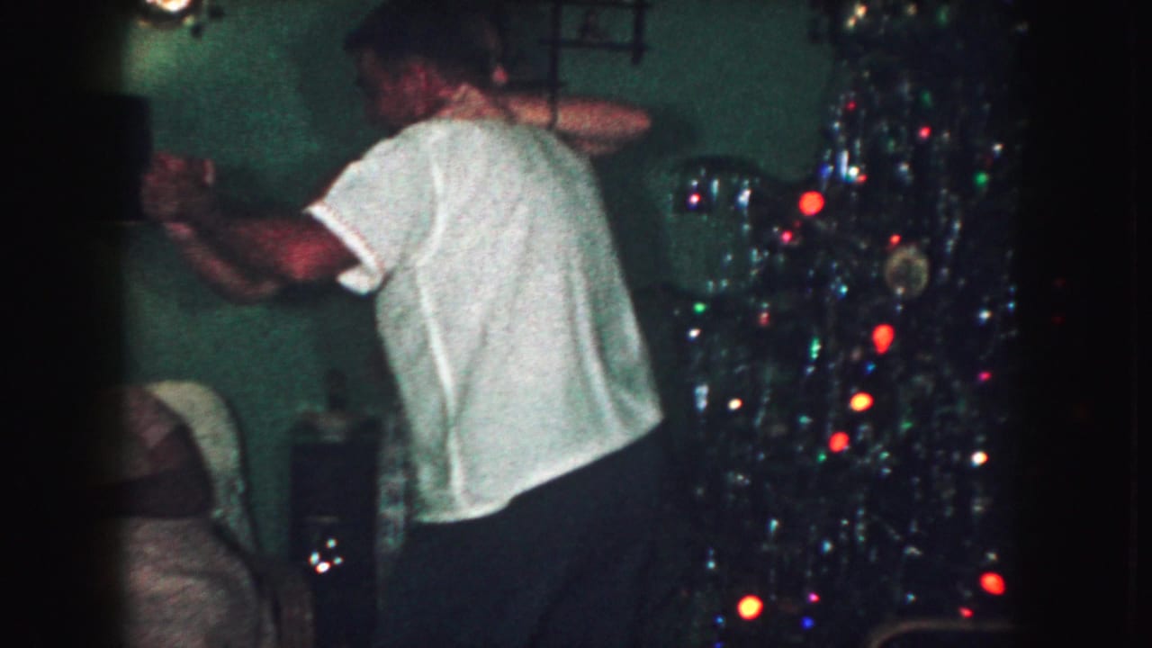 Man in a white shirt dancing near a lit Christmas tree and living-room wall.