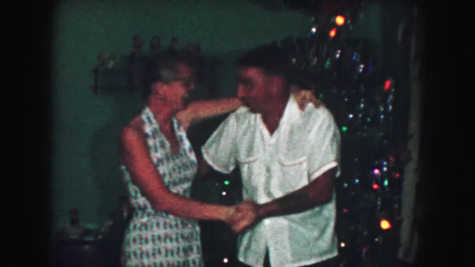An adult couple holds hands and smiles while dancing near a lit Christmas tree in the Kansas City home-movie clip.