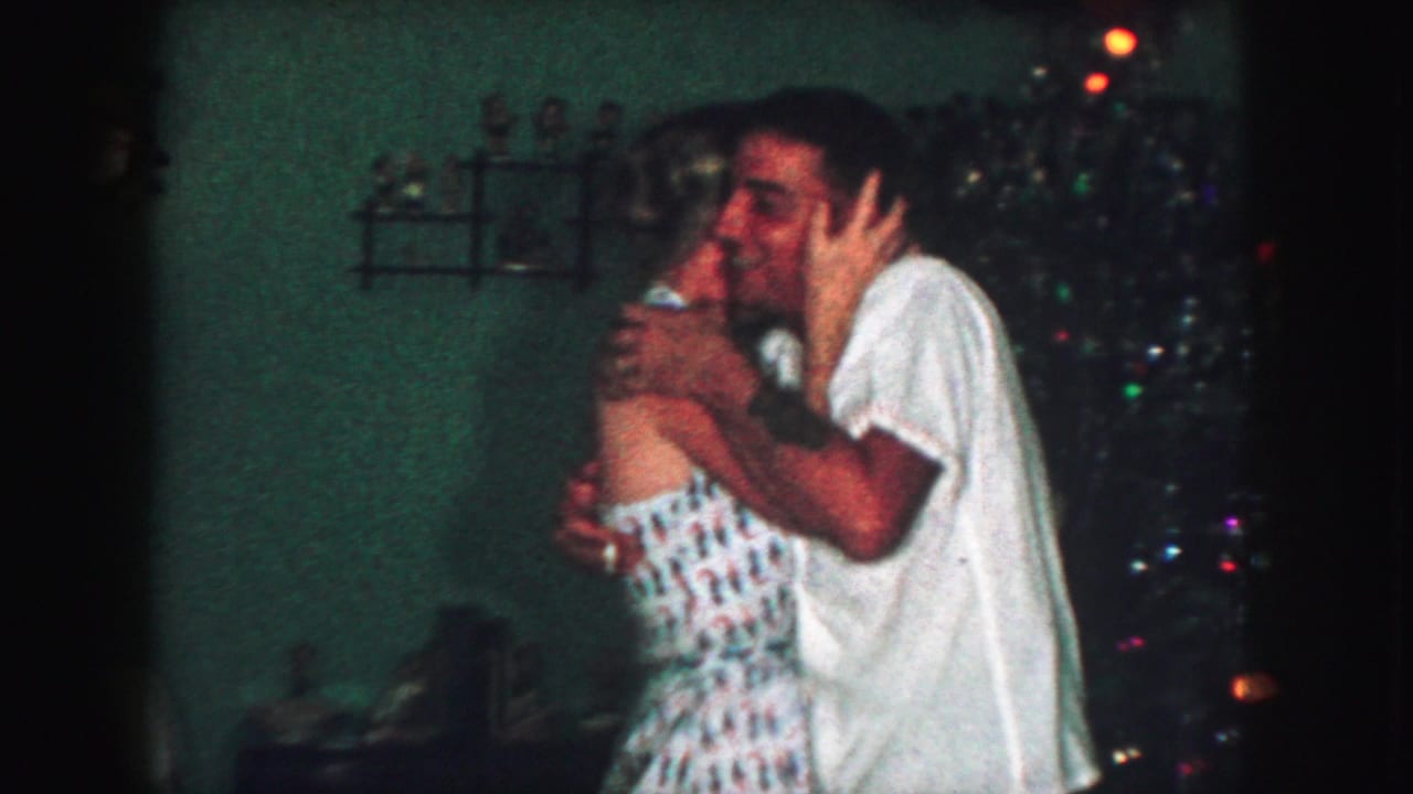 In a 1959 Kansas City home-movie frame, an adult couple embraces and appears to dance beside holiday decorations in a dim living room.