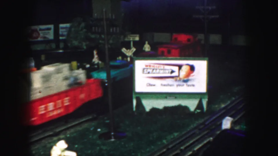 A red model train blurs past a Wrigley's Spearmint sign in a 1959 Kansas City home-movie holiday railroad display.