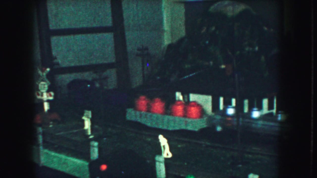 Dim 1959 home-movie frame of a model railroad area with miniature figures, red barrels, trackside posts, and a railroad crossing sign.