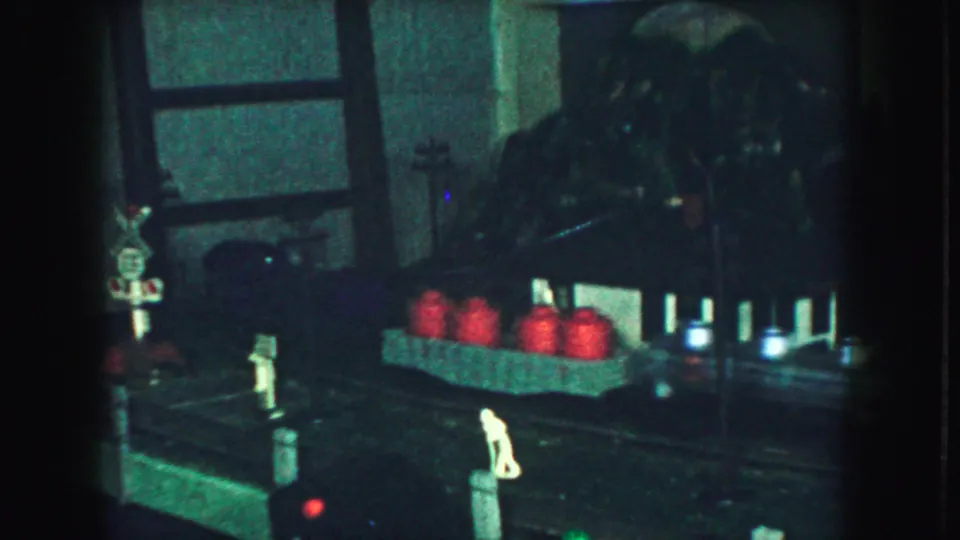 Dim 1959 home-movie frame of a model railroad area with miniature figures, red barrels, trackside posts, and a railroad crossing sign.