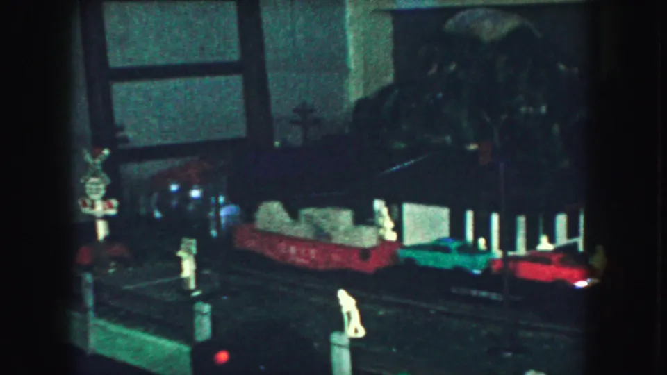 1959 Kansas City home-movie frame showing a toy train layout with a green car, red train cars, miniature figures, and a crossing sign.