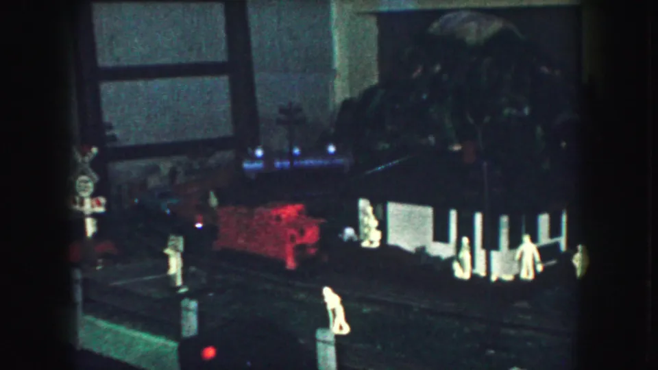 Late frame from the 1959 8mm clip with a red toy train, miniature figures, buildings, and trackside details in a dim model railroad setup.