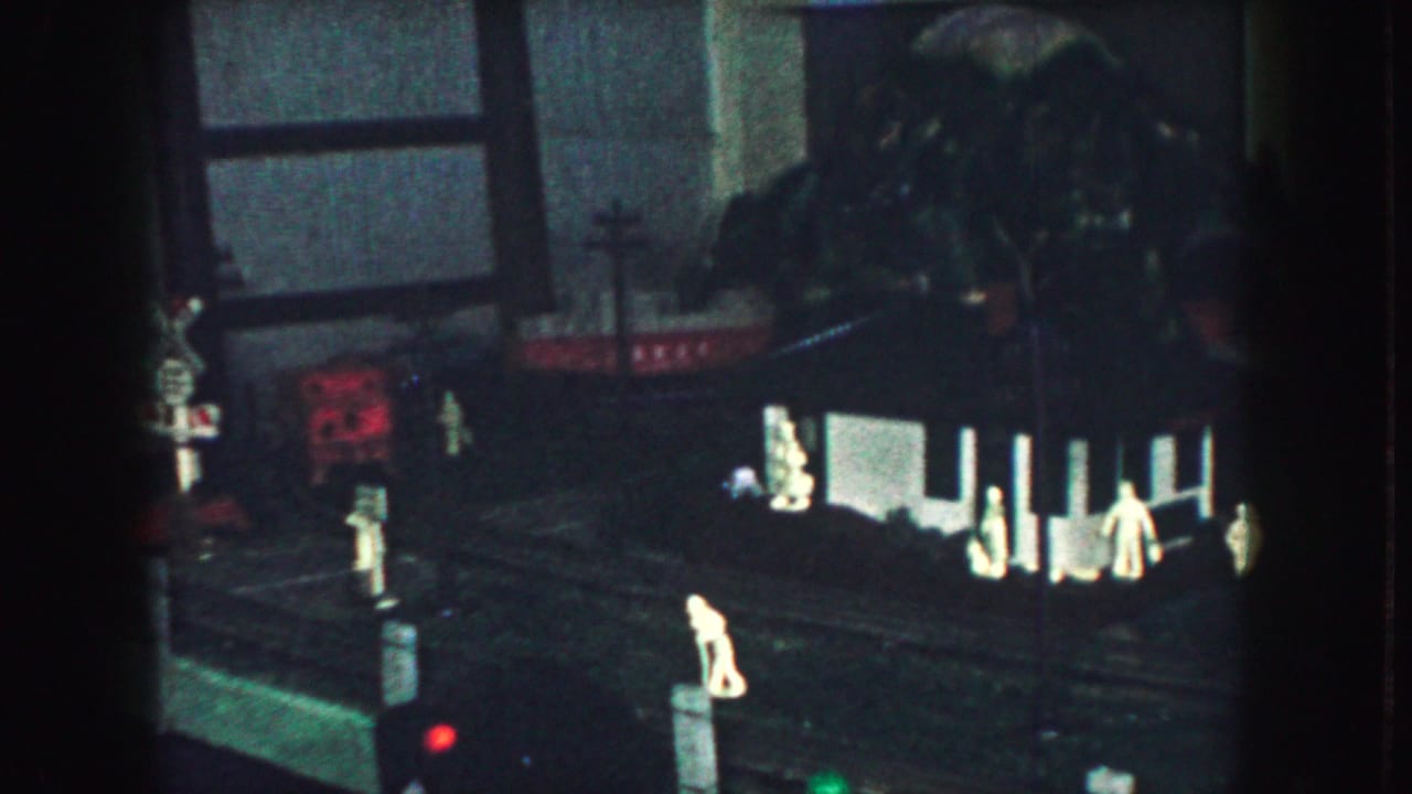 1959 Kansas City home-movie frame showing a dark model train layout with small figures, tracks, and an illuminated miniature structure.