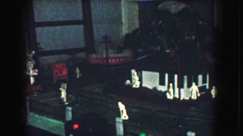 1959 Kansas City home-movie frame showing a dark model train layout with small figures, tracks, and an illuminated miniature structure.