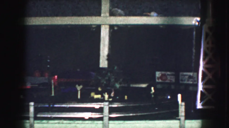 1959 Kansas City home-movie frame with a red model locomotive moving through a dark miniature layout below a pale cross-like support.