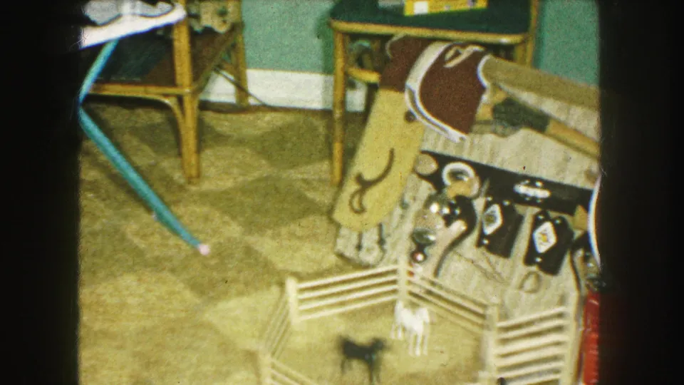1959 Kansas City home-movie frame with toy animals and a small corral below Western accessories, with an ironing board visible nearby.