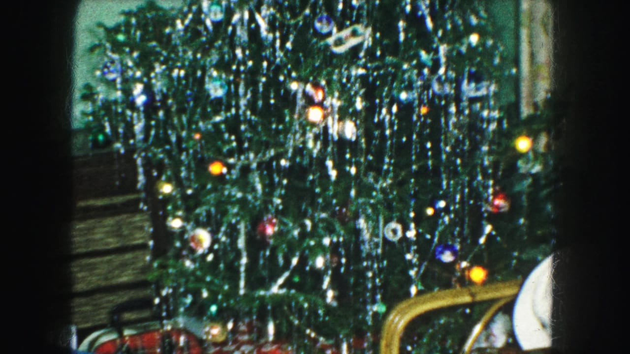 Decorated Christmas tree with silver tinsel, colored ornaments, and lights in a green-walled room.